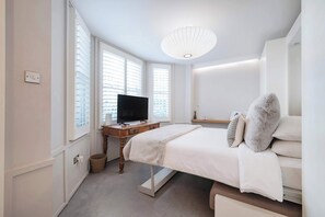 Une chambre à coucher avec un lit, un téléviseur posé sur une table en bois et une grande fenêtre avec des stores.