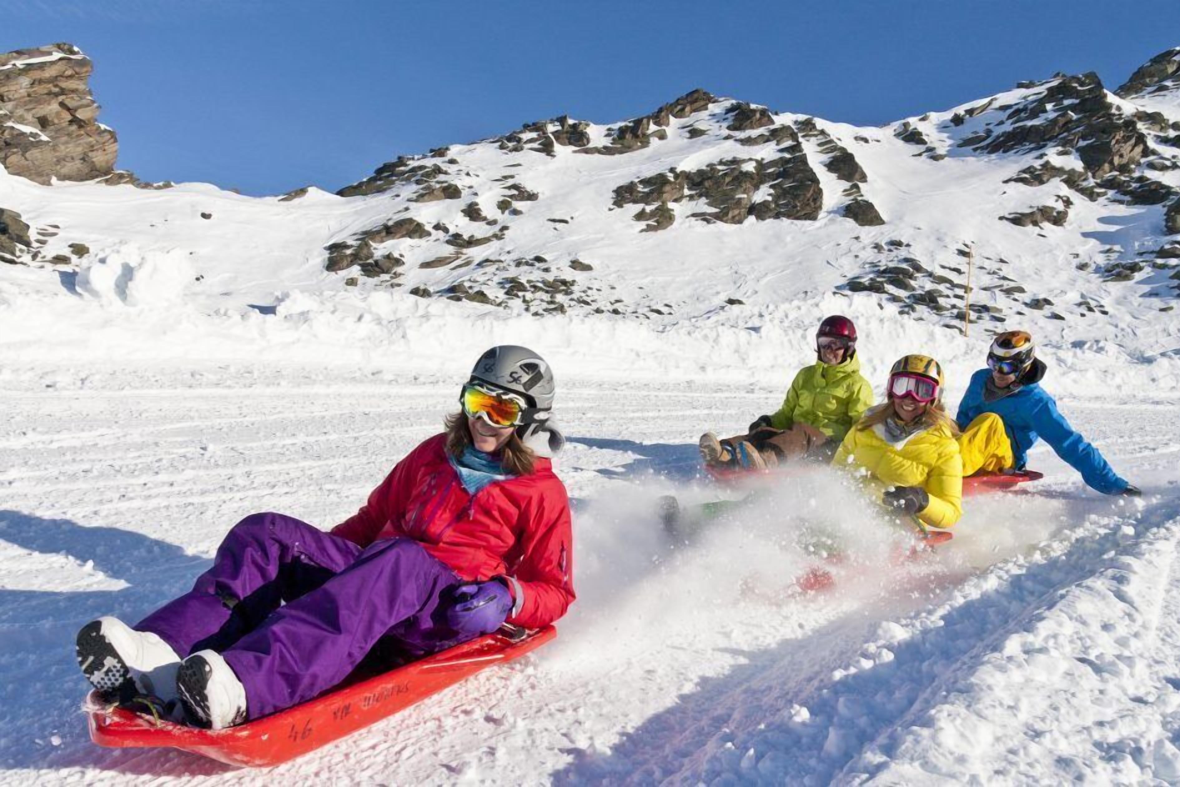 Four people sledding down a snowy slope with rocky mountains in the background.