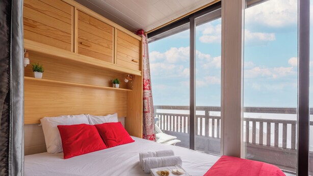 A hotel room with a large bed, wooden headboard, a view of the balcony, and a window with a curtain. - Cimes des caron  (Les Belleville)