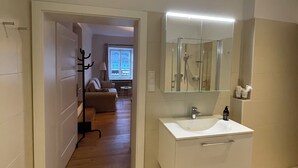 A modern bathroom with a white sink, mirror, and shower. There is a purple rug on the floor and a hallway leading to a living area. - Gästehaus Burgblick im Weinhaus Pachler (Hochburg-Ach)