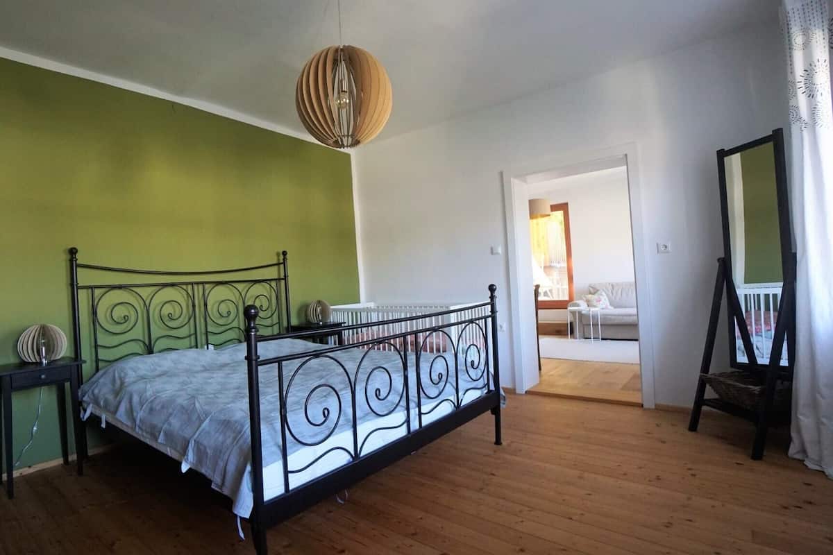 Ein Schlafzimmer mit einem Metallbettgestell, einer grünen Wand, einem Holzboden, einer Pendelleuchte und Blick auf einen Balkon mit Stuhl und Tisch.
