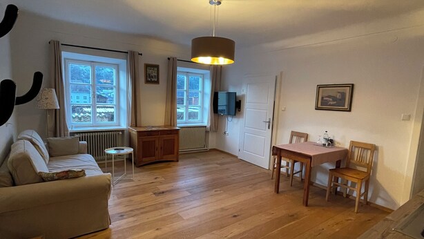 A spacious living area with a wooden floor, a sofa, a dining table with chairs, a ceiling light, and a window with curtains. - Gästehaus Burgblick im Weinhaus Pachler (Hochburg-Ach)