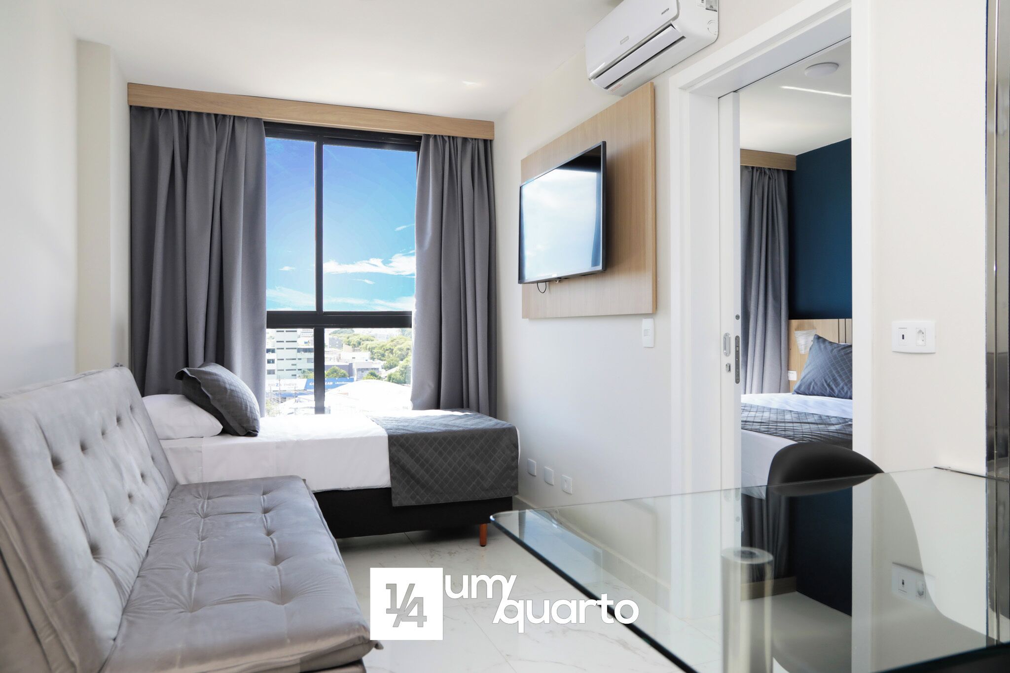 A modern hotel room with a bed, a sofa, a TV, and a glass table.