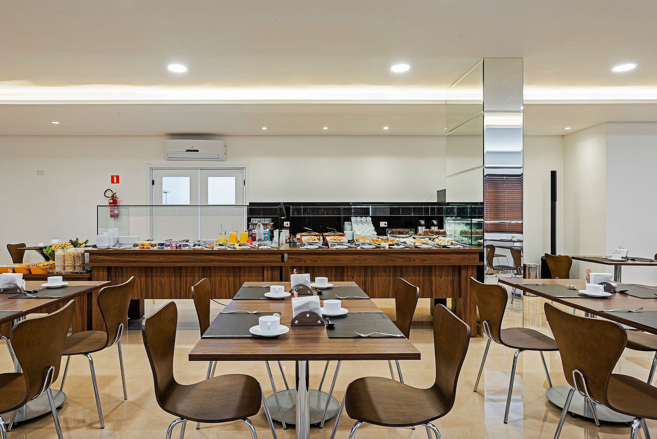 Een moderne eetruimte met meerdere tafels en stoelen, een buffet in de achtergrond, en een brandblusser aan de muur.