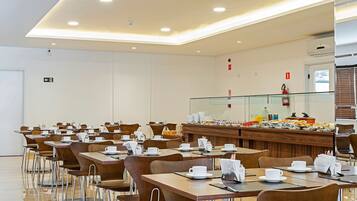 Un comedor de hotel con varias mesas y sillas, una barra de buffet con distintos alimentos y una división de vidrio.