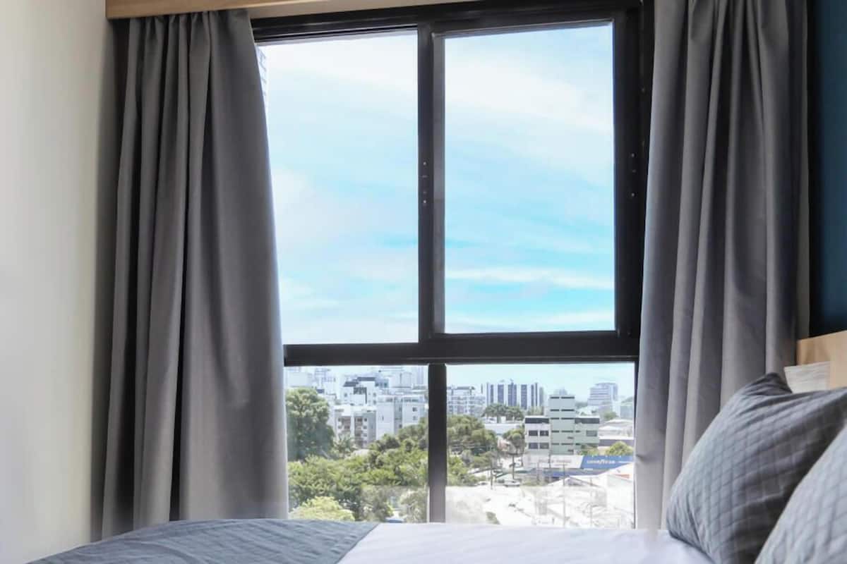 A hotel room with a large window, a bed with grey bedding, and a view of a cityscape.