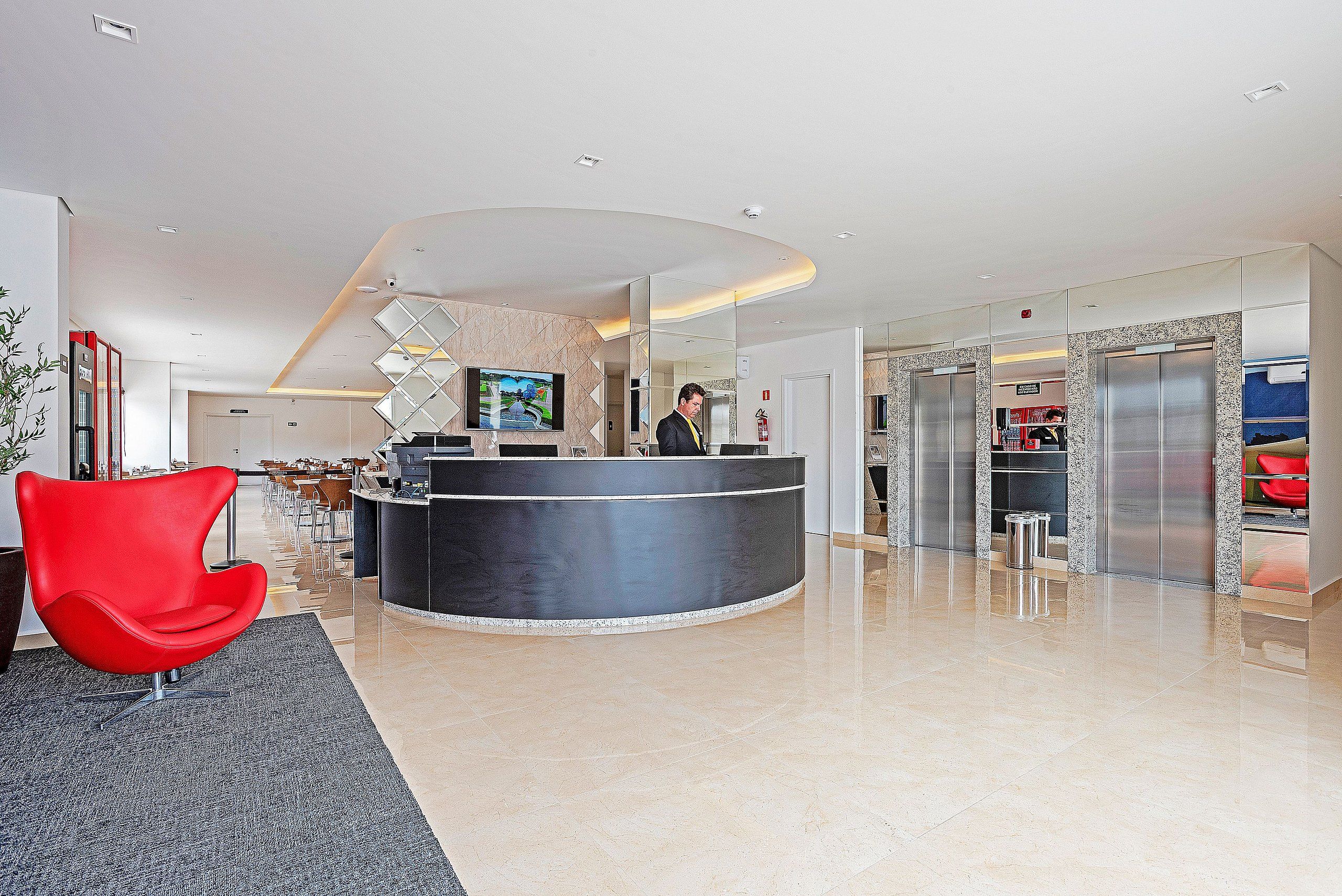 A modern hotel lobby with a reception desk, a red chair, and a large TV screen.