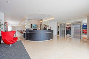 Um hall de hotel moderno com uma mesa de recepção, uma cadeira vermelha e uma grande tela de televisão.