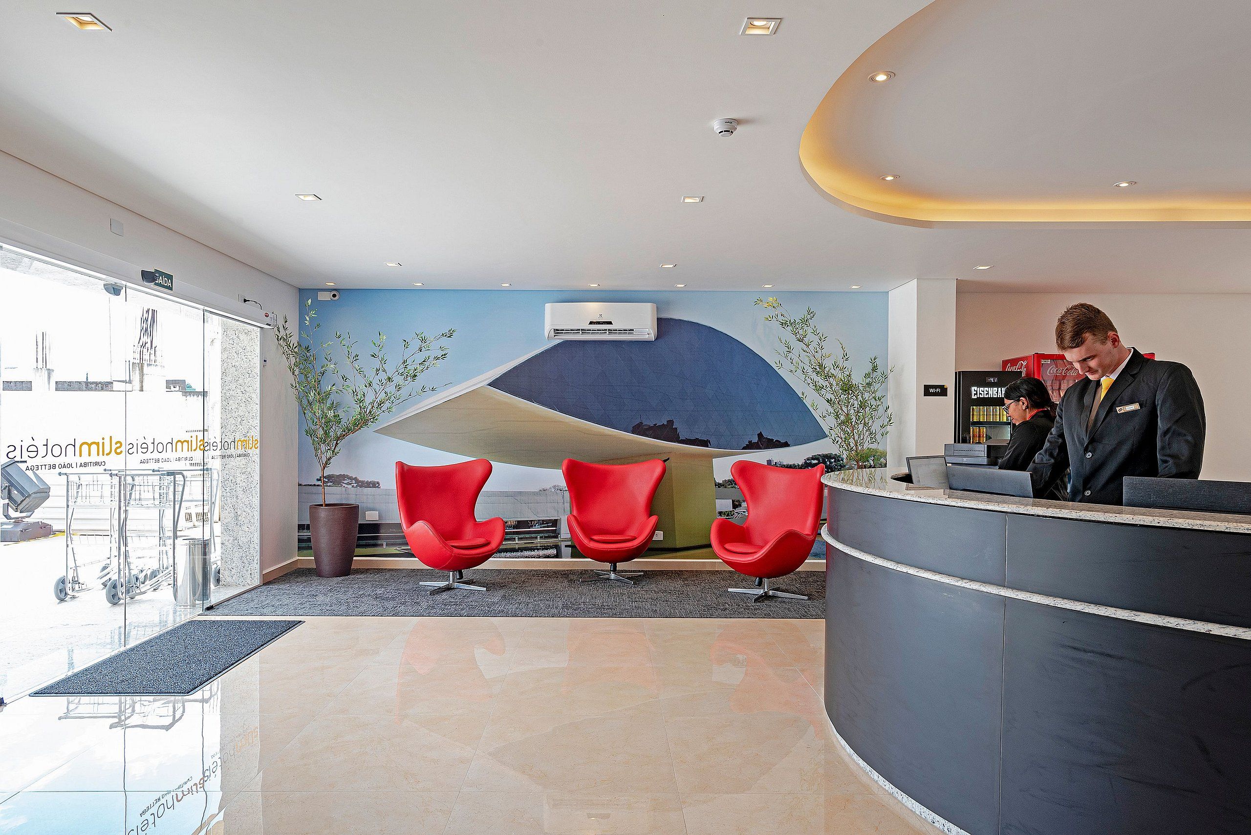 Ein Hotel-Empfangsbereich mit einer gebogenen Rezeption, roten Sesseln, einem großen Wandbild und einem modernen Deckenstil.