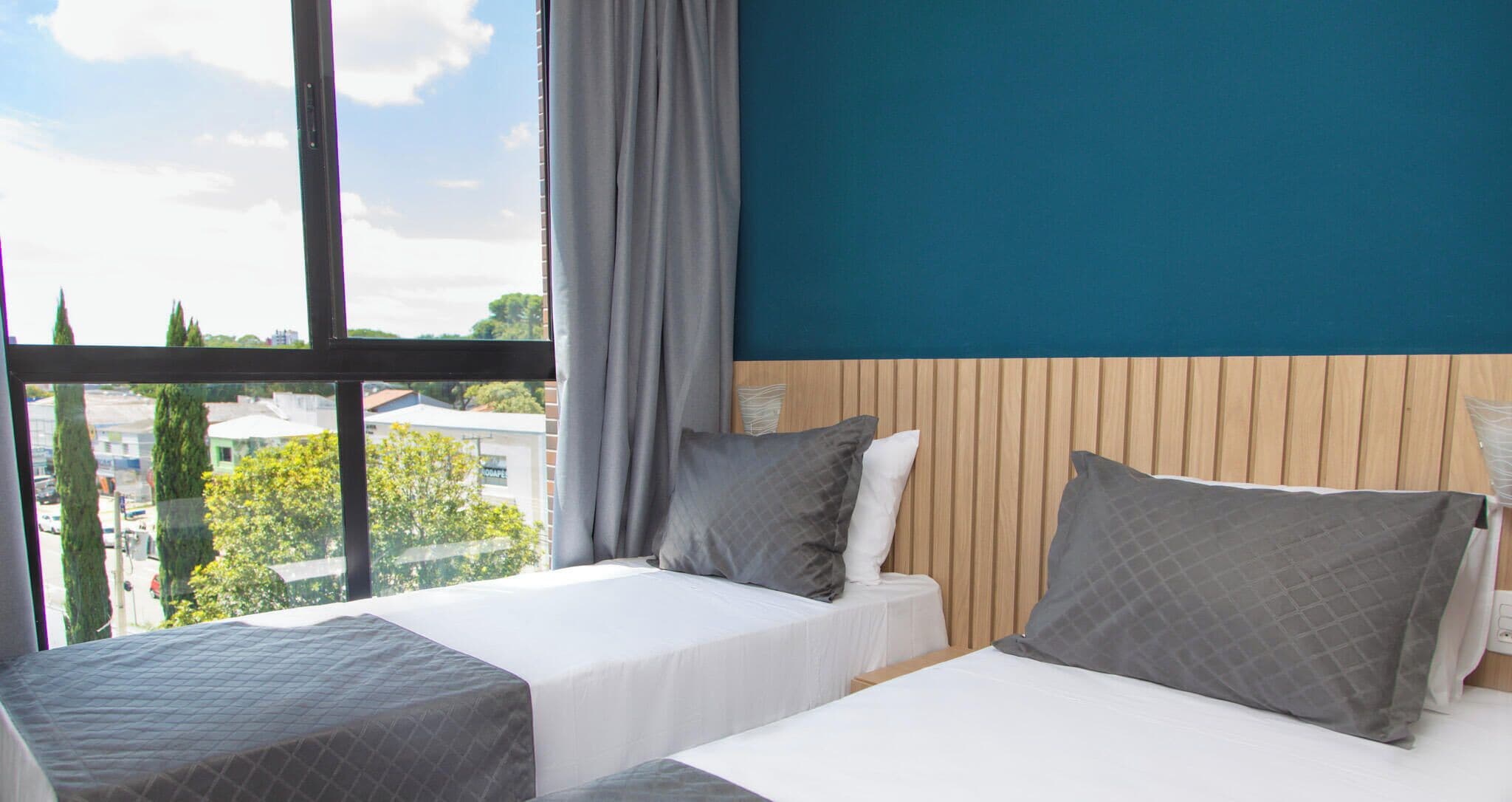 Dos camas individuales con ropa de cama gris en una habitación con un ventanal grande, paneles de madera en la pared y vistas a edificios y árboles.