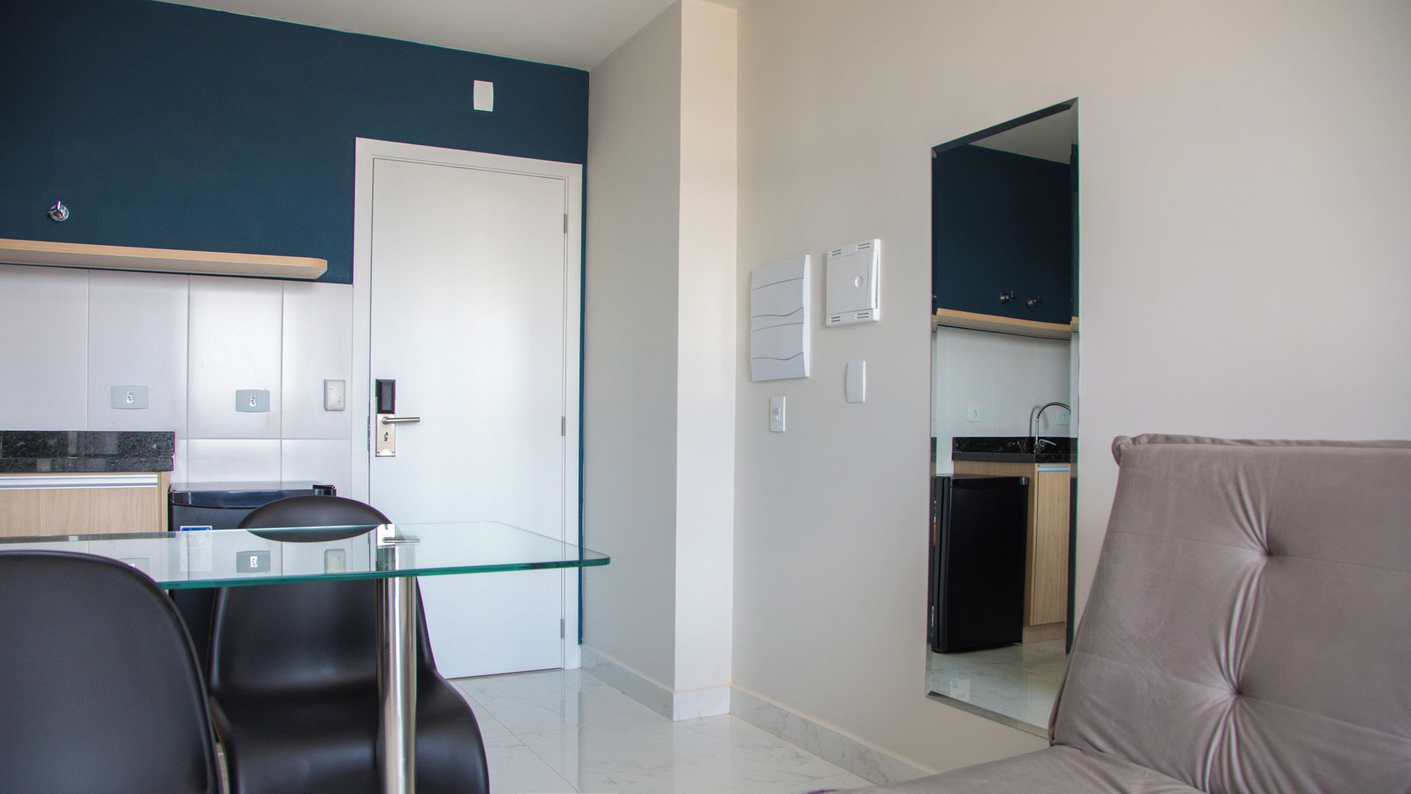 Uma sala de estar moderna com uma mesa de jantar de vidro, cadeiras pretas e um sofá bege. Uma cozinha visível ao fundo com uma porta branca.
