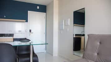 A modern living room with a glass dining table, black chairs, and a beige sofa. A kitchen is visible in the background with a white door.