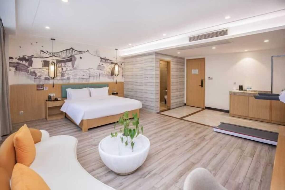 Ein modernes Hotelzimmer mit einem Bett, einem Schreibtisch, einem Sessel, einer Couch, einem Wandbild und einer Pflanze.