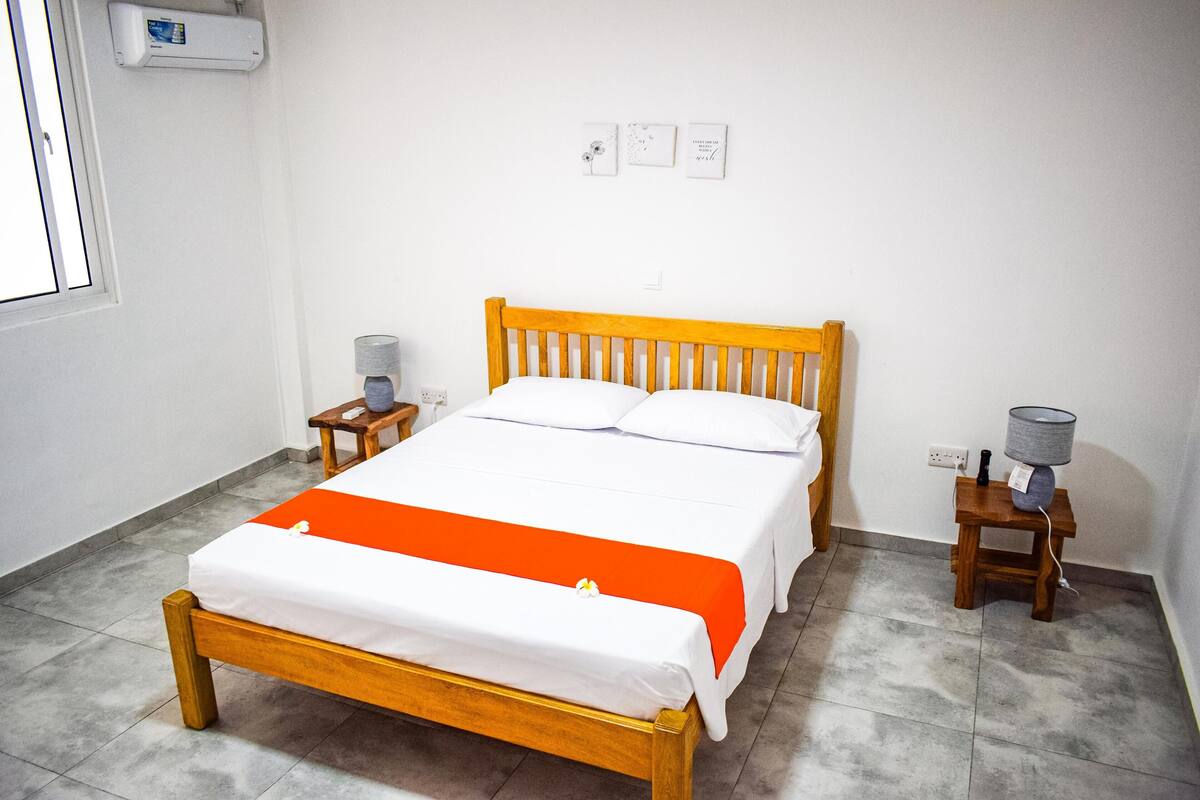 A neatly made bed with a wooden frame and white bedding, a small bedside table with a lamp, and a wall-mounted air conditioner.
