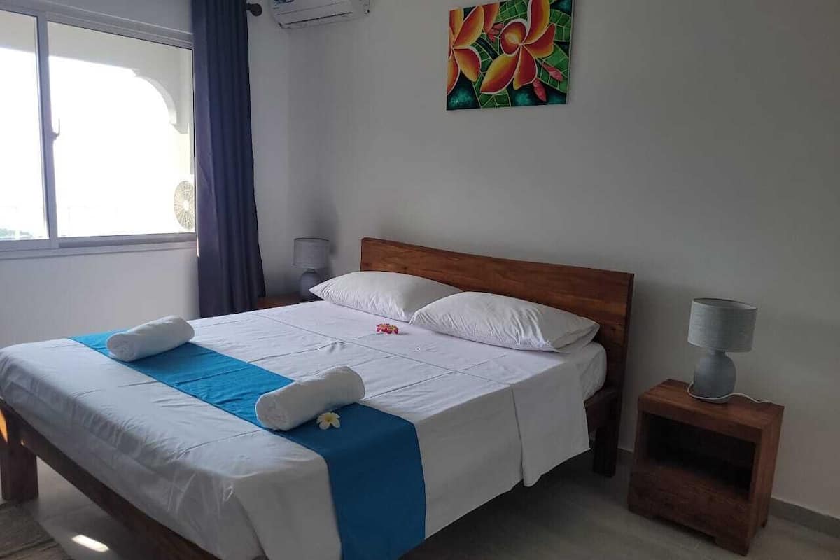 A neatly made bed with white linens and a blue accent, a wooden headboard, a small bedside table with a lamp, and a colorful painting on the wall.