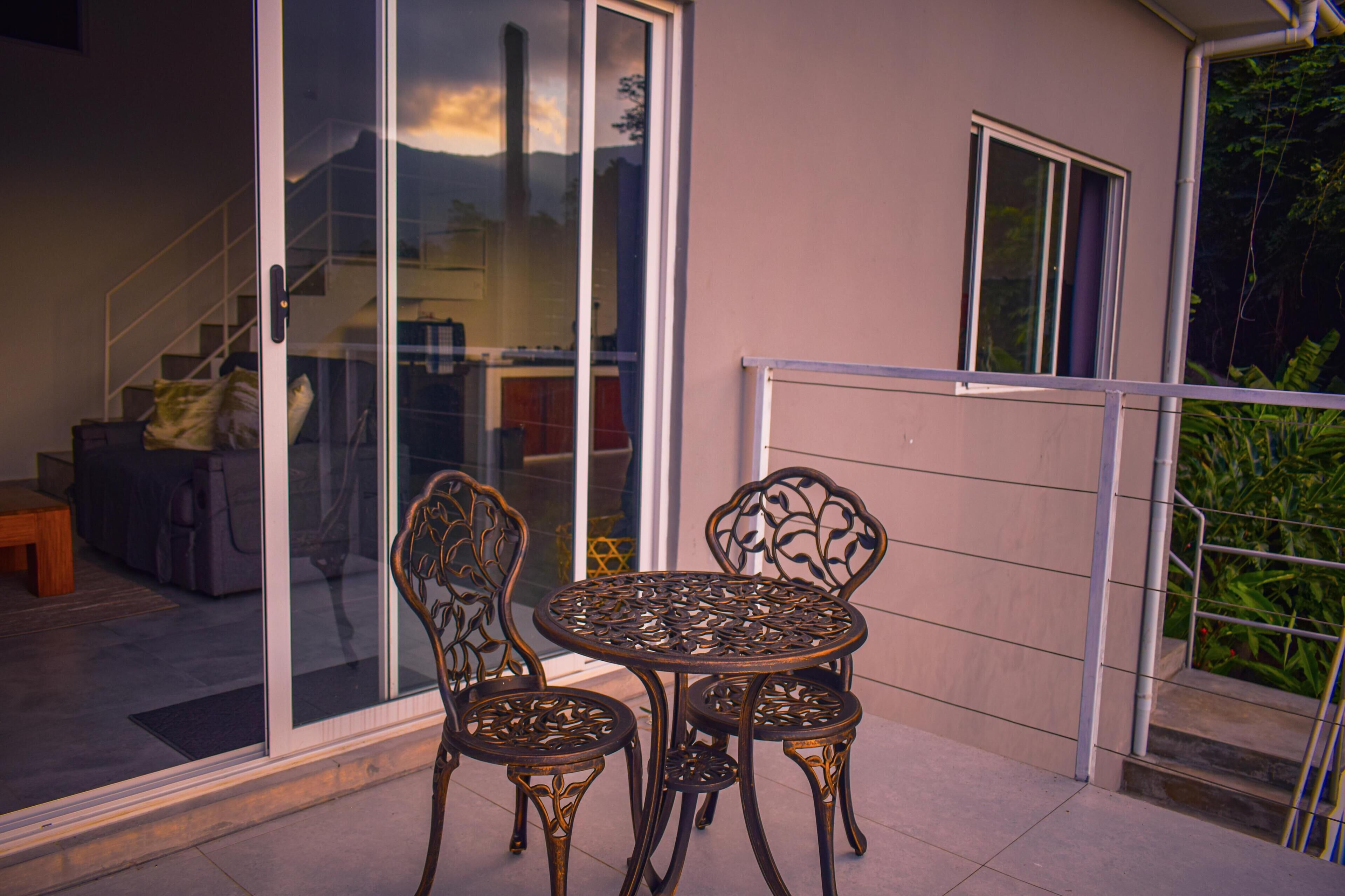 A patio with a glass door leading to a staircase, a round metal table with chairs, and a view of a building with a window and a door.