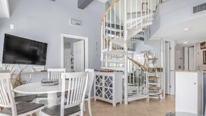 A spacious living area with a white staircase, a dining table with chairs, and a flat-screen TV mounted on the wall. - GS Plantation West 1339 (Gulf Shores)