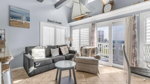 A living room with a gray sofa, a round glass coffee table, and a chair. There are two windows with white shutters, a sailboat decoration, and a view of a building outside. - GS Plantation West 1339 (Gulf Shores)