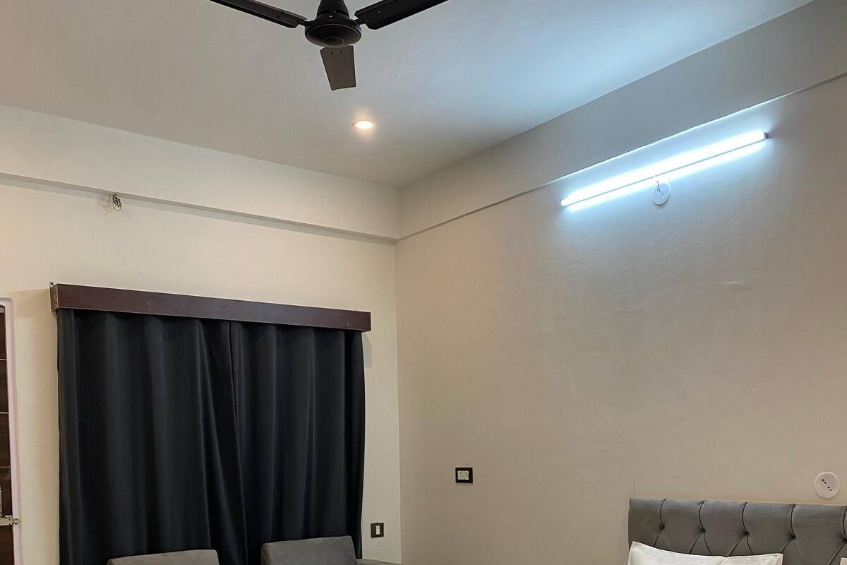 A hotel room with a bed, two armchairs, a small table, a ceiling fan, and a window with curtains.