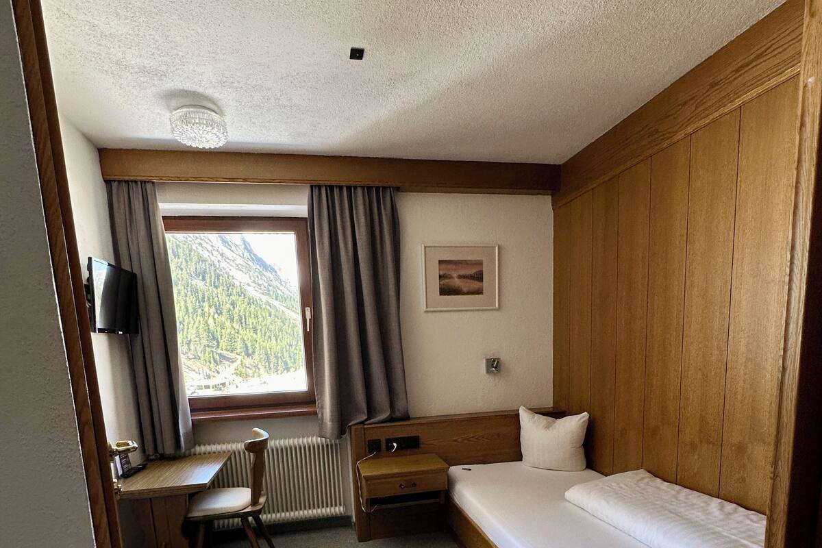 A hotel room with a wooden bed, a desk with a chair, a radiator, a window with curtains, and a picture on the wall.