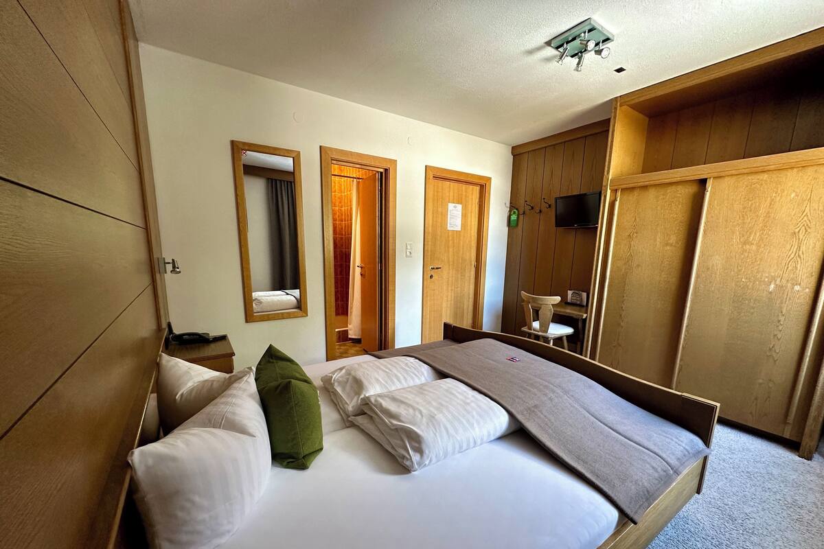 A hotel room with a bed, wooden wardrobe, a desk with a chair, and a television.