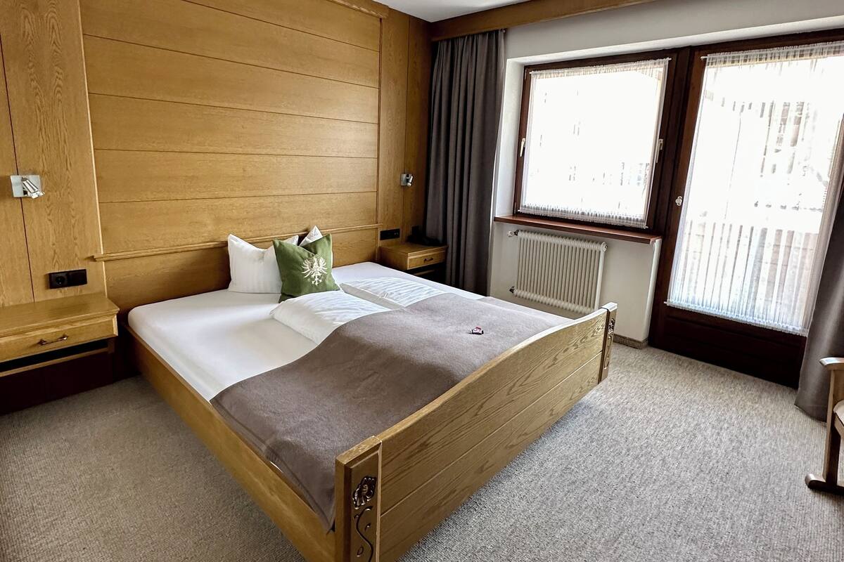 A hotel room with a wooden bed, a nightstand, a window with curtains, and a radiator.