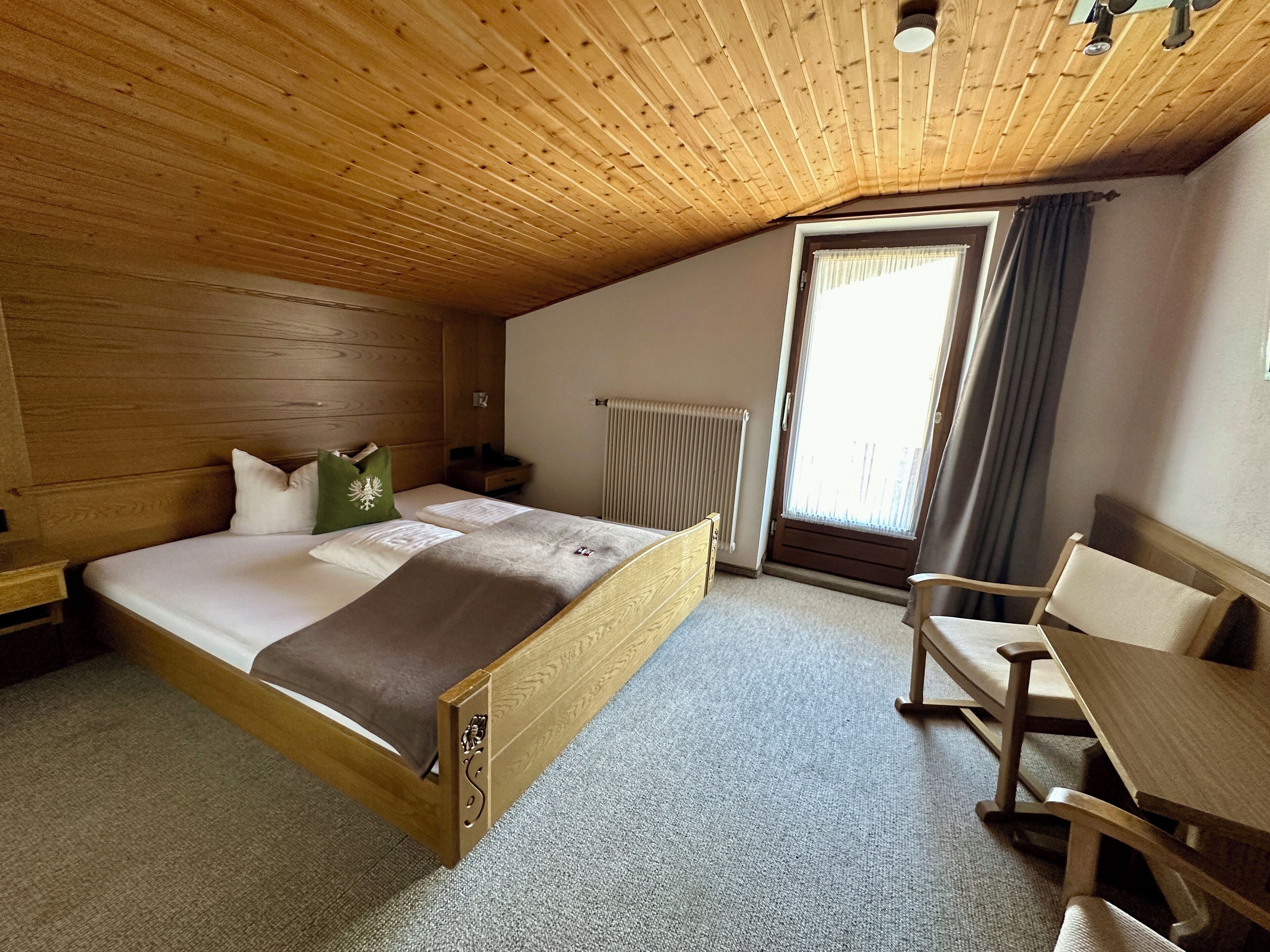 Un dormitorio con techo de madera, una cama con cabecera, una mesita de noche, una silla y una ventana con cortinas.