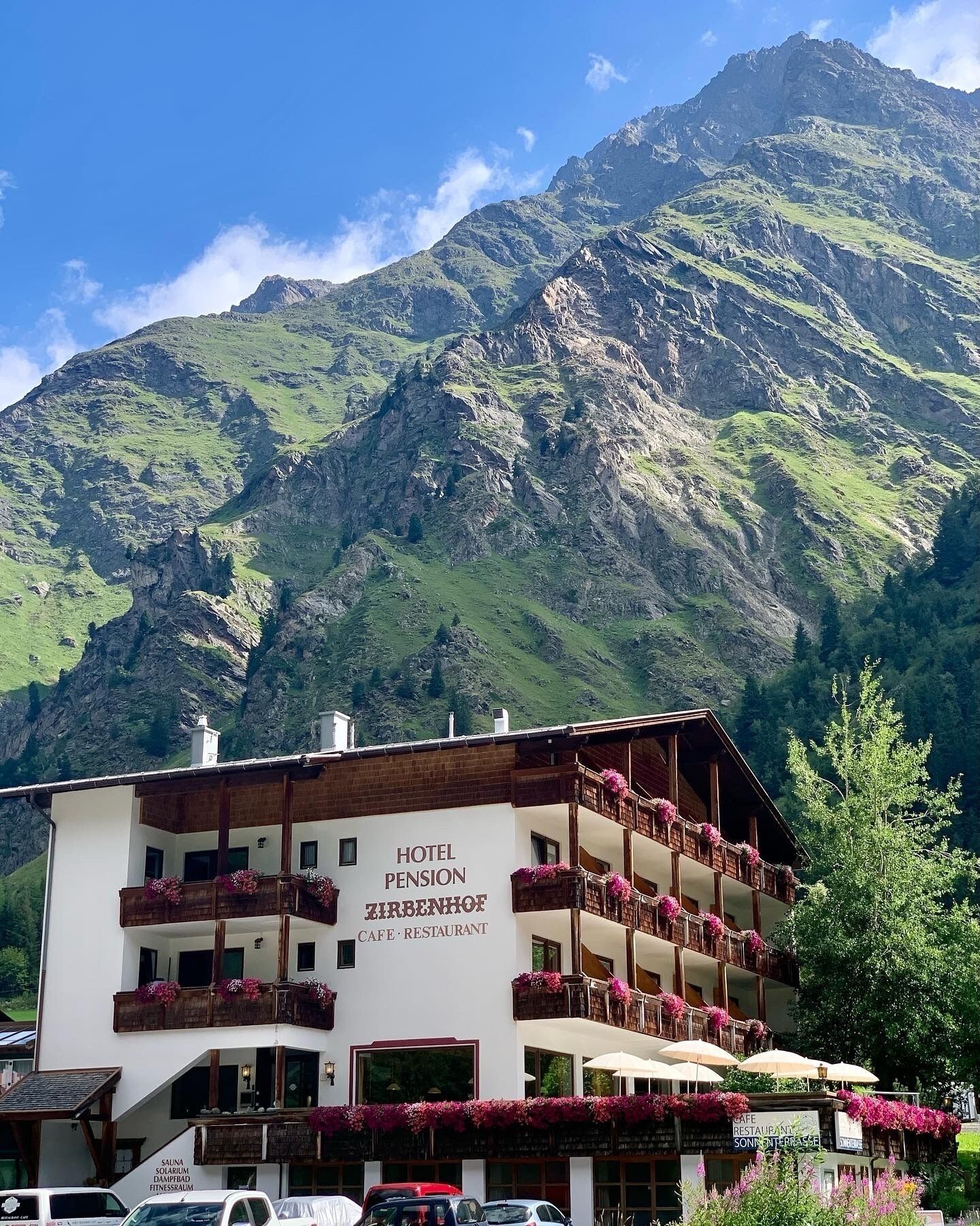 A hotel with multiple balconies adorned with flowers, a sign indicating it is a pension, and a mountain backdrop.