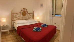 A single bed with a red bedspread and two folded towels, a bedside table with a lamp, and a window with blinds. - Apartment 2-Villa Augusta-del-Sol  (Fontane Bianche)