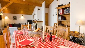 Een eethoek met een rood-wit geruit tafelkleed, houten stoelen, een mand met brood, flessen wijn en een boekenkast met boeken en een klok.