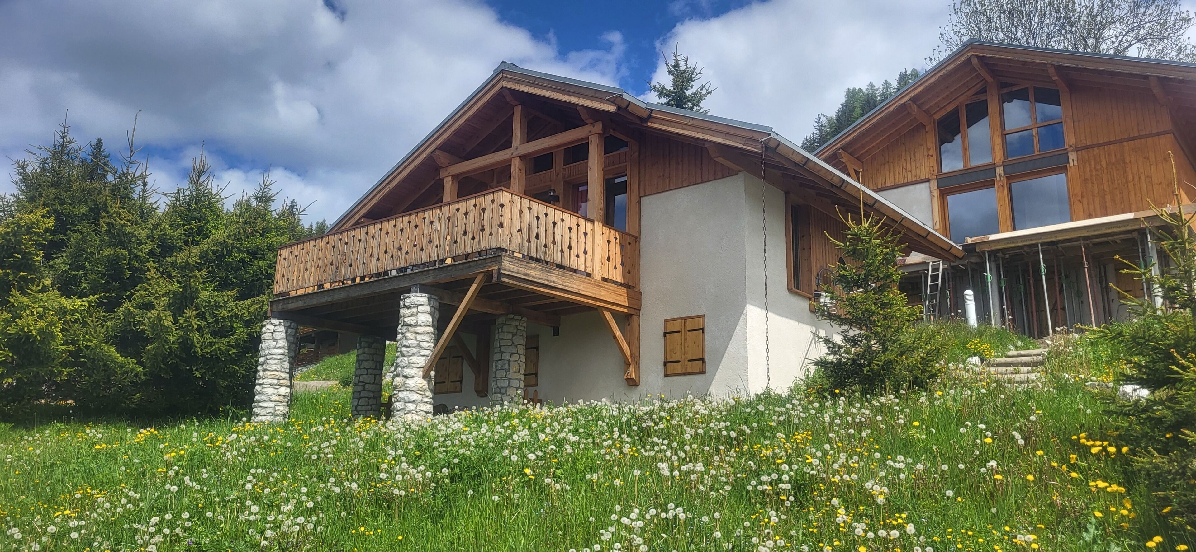 A wooden chalet with a balcony, stone pillars, and a garden with wildflowers.