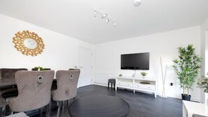 Ein modernes Wohnzimmer mit einem runden schwarzen Tisch, grauen Polsterstühlen, einer weißen Kommode, einem Flachbildfernseher und einer Topfpflanze.