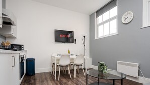 A modern kitchen with a microwave, stove, and cabinets. A dining area with a table and chairs is visible. A TV mounted on the wall displays "NETFLIX".