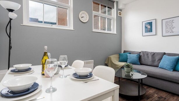 A modern living room with a dining area, grey walls, a grey sofa with blue cushions, a round coffee table, and a dining table set with plates, glasses, and a bottle of wine.