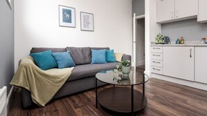 A modern living room with a grey sofa, teal cushions, a glass coffee table, and a kitchen area with white cabinets and a countertop.