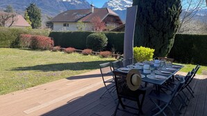 Terraza con una mesa lista para comer, rodeada de un jardín cuidado y vistas a la montaña.