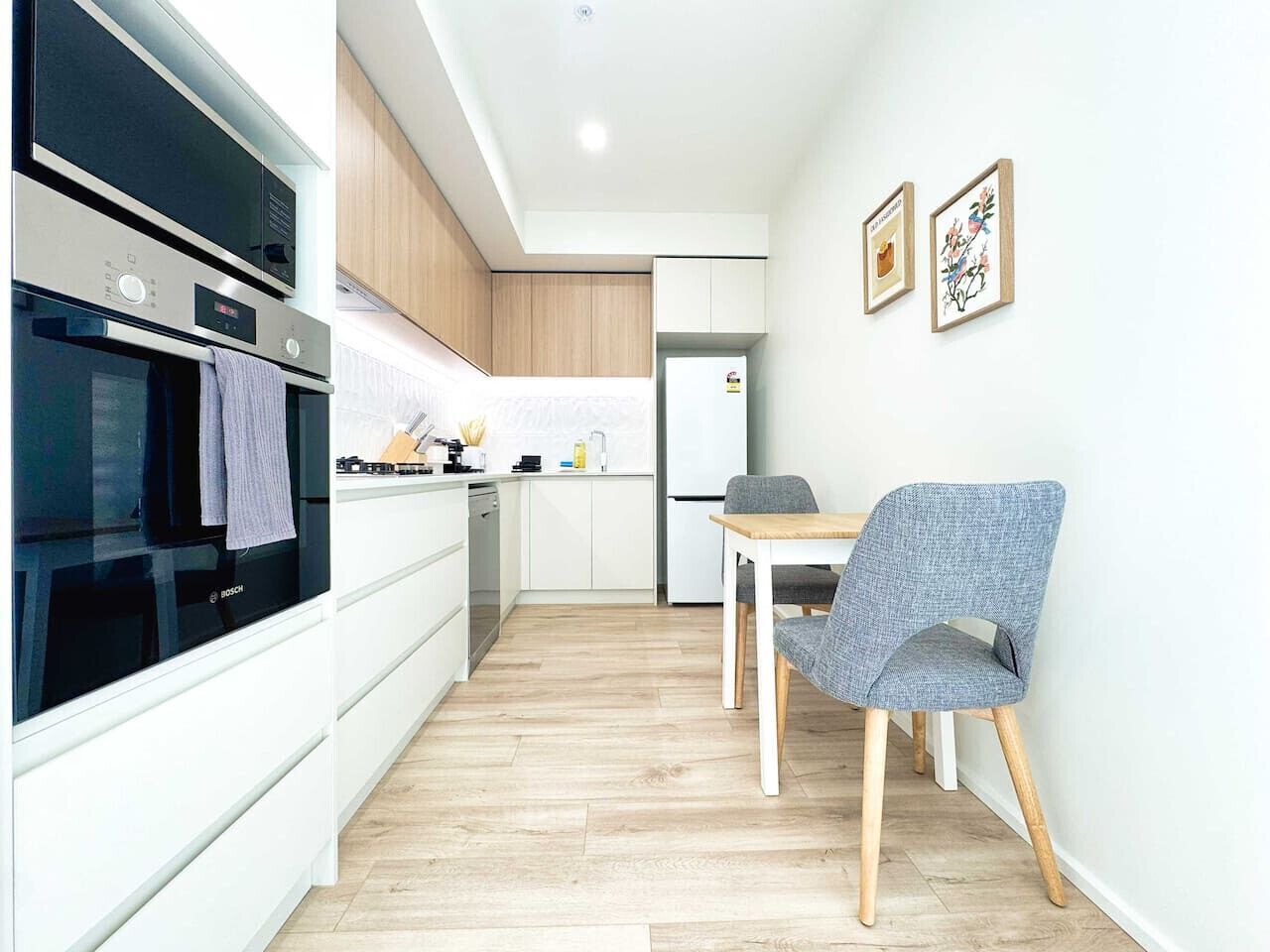 A modern kitchen with white cabinets, a Bosch oven, a dining table with a chair, and two framed pictures on the wall.