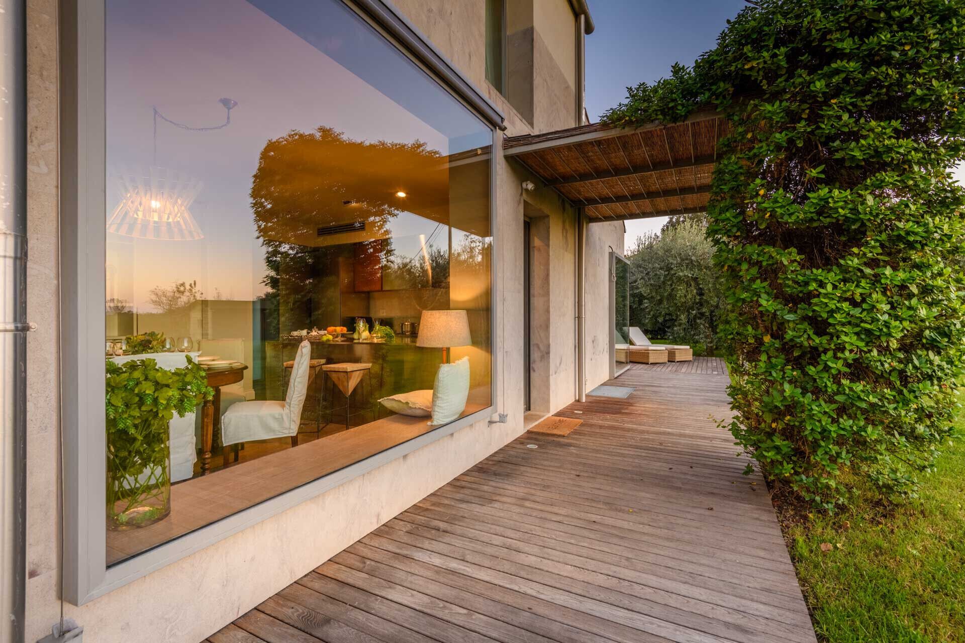 A modern outdoor dining area with a wooden deck, glass walls, and a view of the interior.