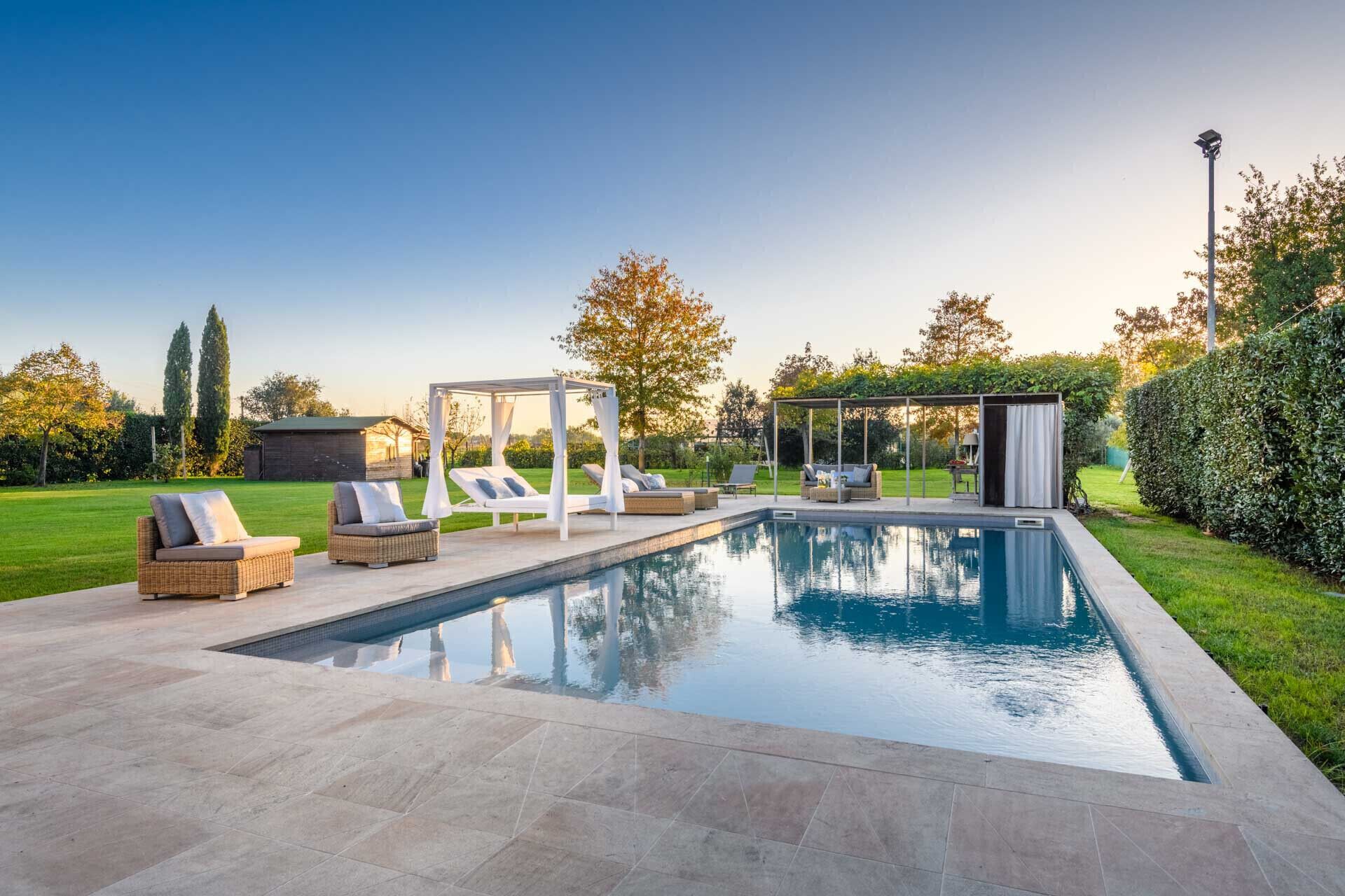 A rectangular swimming pool with a tiled deck, surrounded by lounge chairs and a covered seating area.