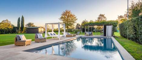 A rectangular swimming pool with a tiled deck, surrounded by lounge chairs and a covered seating area.