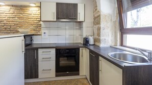 Een kleine keuken met witte kastjes, een inbouwoven, een accentmuur van steen en een raam dat voor daglicht zorgt.