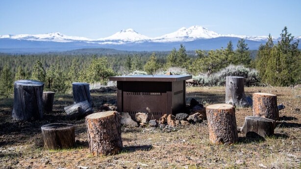 Un décor extérieur rustique, avec une structure métallique entourée de souches d’arbres, le tout se dressant sur fond de montagnes enneigées et d’une forêt dense.