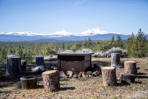 A rustic outdoor setting with a metal structure surrounded by tree stumps, set against a backdrop of snow-capped mountains and dense forest. - Vista View Retreat – Serene Mountain Escape (Sisters)