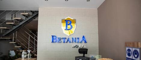 A hotel reception area with a modern staircase, a reception desk, and a logo for 'Betania Hotel'.