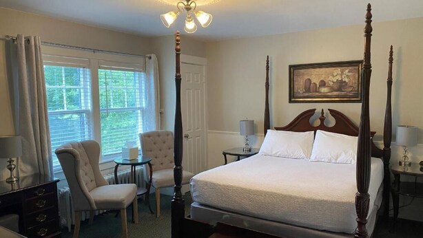 A four-poster bed with a white bedspread, a chair, a small table, and a lamp.