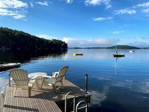 Un muelle con sillas y una mesa, agua tranquila y barcos a lo lejos.