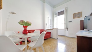 Un comedor con una mesa redonda, sillas blancas, un jarrón rojo con flores y un sofá rojo.