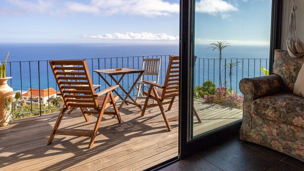 Eine Holzterrasse mit Tisch und Stühlen, mit Blick auf eine Küstenlandschaft, in der Ferne Meer und Berge.