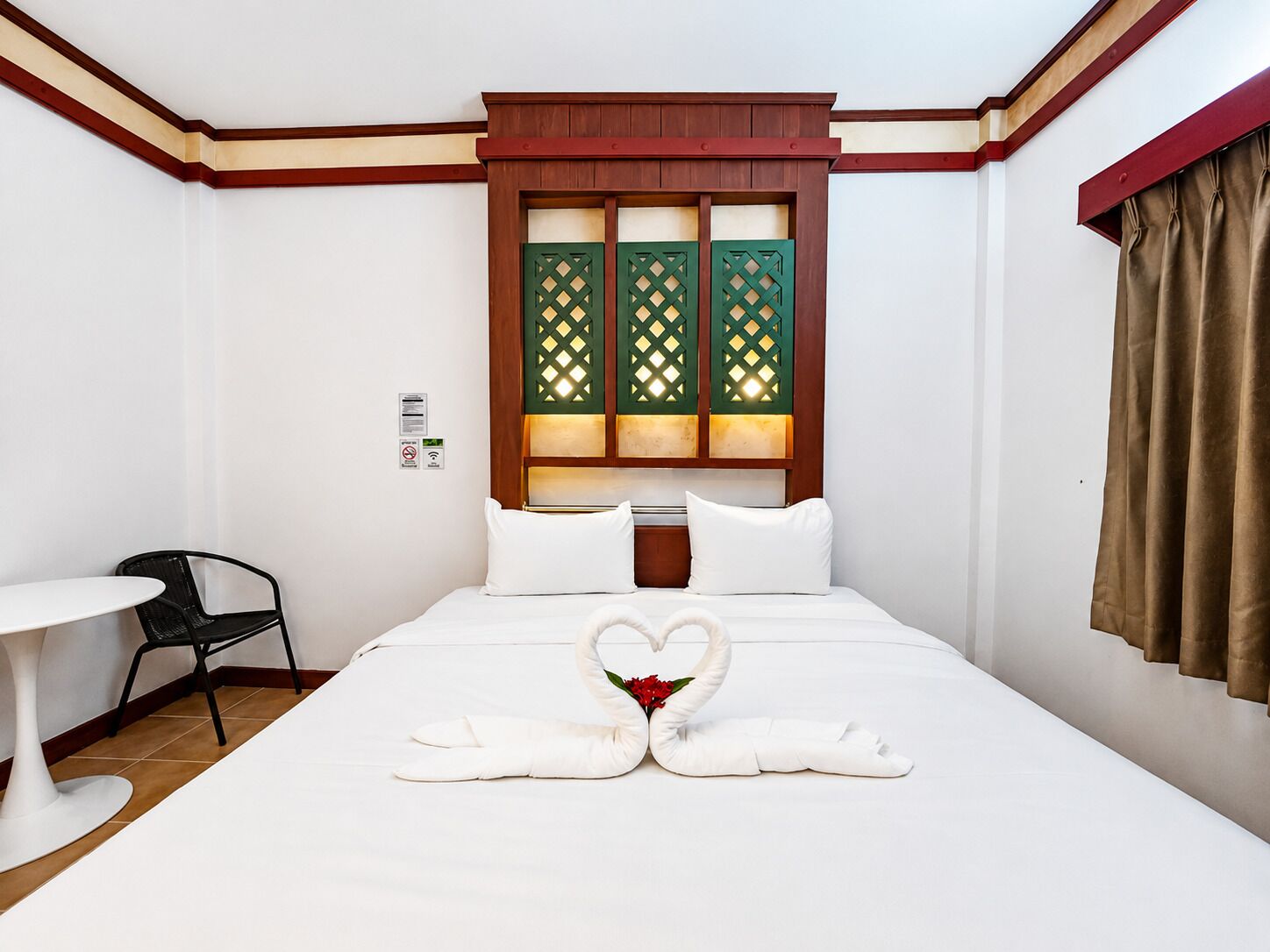 Una habitación de hotel con una cama, una mesita pequeña, una silla y una ventana con marco de madera y paneles de celosía verde.