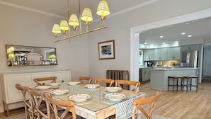 Un coin repas avec une table en verre entourée de chaises en bois, un lustre et une cuisine en arrière-plan.